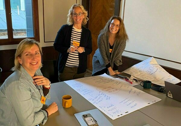 Three smiling women stand around a table with large white papers covered in blue handwritten brainstorming notes, diagrams, and circled words like "HOPE."